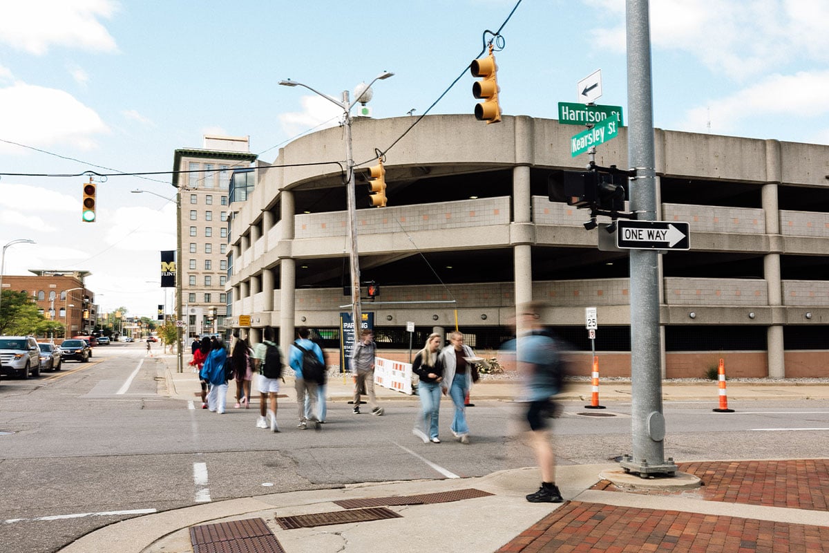 FLINT_First Day of Class Fall 2026_0042 A busy street scene showing pedestrians crossing in front of a parking garage, with traffic lights and street signs visible.