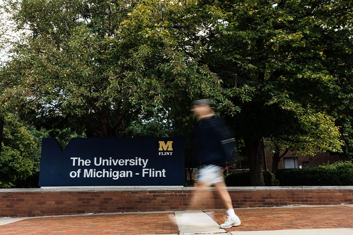 FLINT_First Day of Class Fall 2026_0043 A person walking past a sign that reads "The University of Michigan - Flint" surrounded by trees.