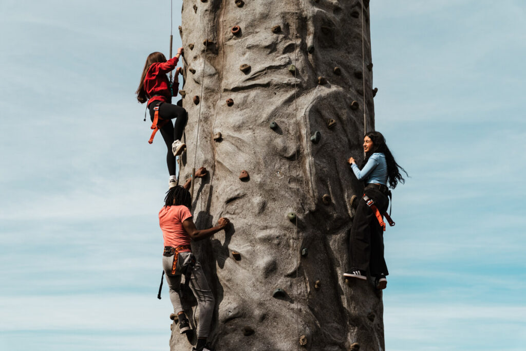 Three individuals are climbing a rock wall outdoors. One person is near the top, while the others are lower down, all wearing harnesses.