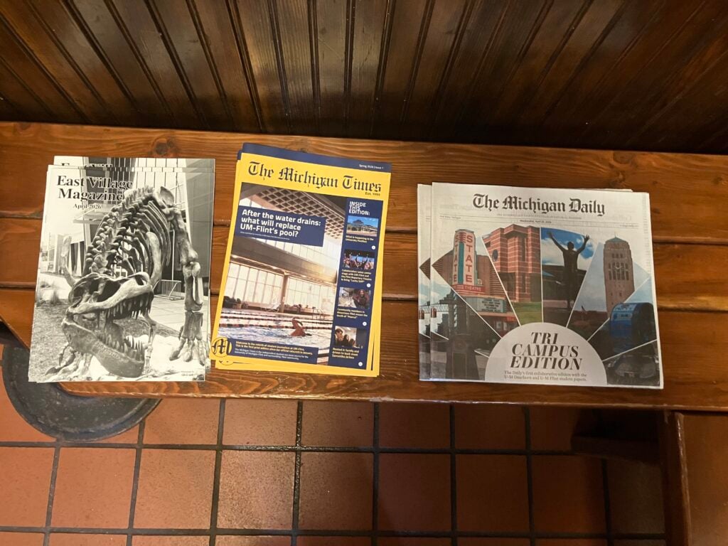 Three magazines are displayed on a wooden bench: "East Village Magazine" featuring a dinosaur skeleton, "The Michigan Times" with an article about a pool replacement, and "The Michigan Daily" labeled "TRI CAMPUS EDITION" with a collage of local buildings.