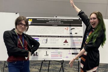 A professor and a student stand on either side of a research poster displayed on an easel at a conference. The poster displays charts, diagrams, and molecular illustrations related to bacterial antibiotic sensitivity research. The student on the right raises one arm and smiles broadly, while the professor on the left stands with arms crossed, smiling.