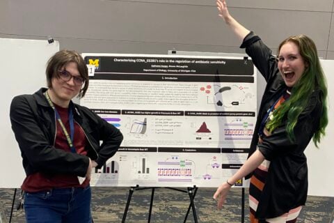 A professor and a student stand on either side of a research poster displayed on an easel at a conference. The poster displays charts, diagrams, and molecular illustrations related to bacterial antibiotic sensitivity research. The student on the right raises one arm and smiles broadly, while the professor on the left stands with arms crossed, smiling.