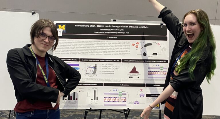 A professor and a student stand on either side of a research poster displayed on an easel at a conference. The poster displays charts, diagrams, and molecular illustrations related to bacterial antibiotic sensitivity research. The student on the right raises one arm and smiles broadly, while the professor on the left stands with arms crossed, smiling.