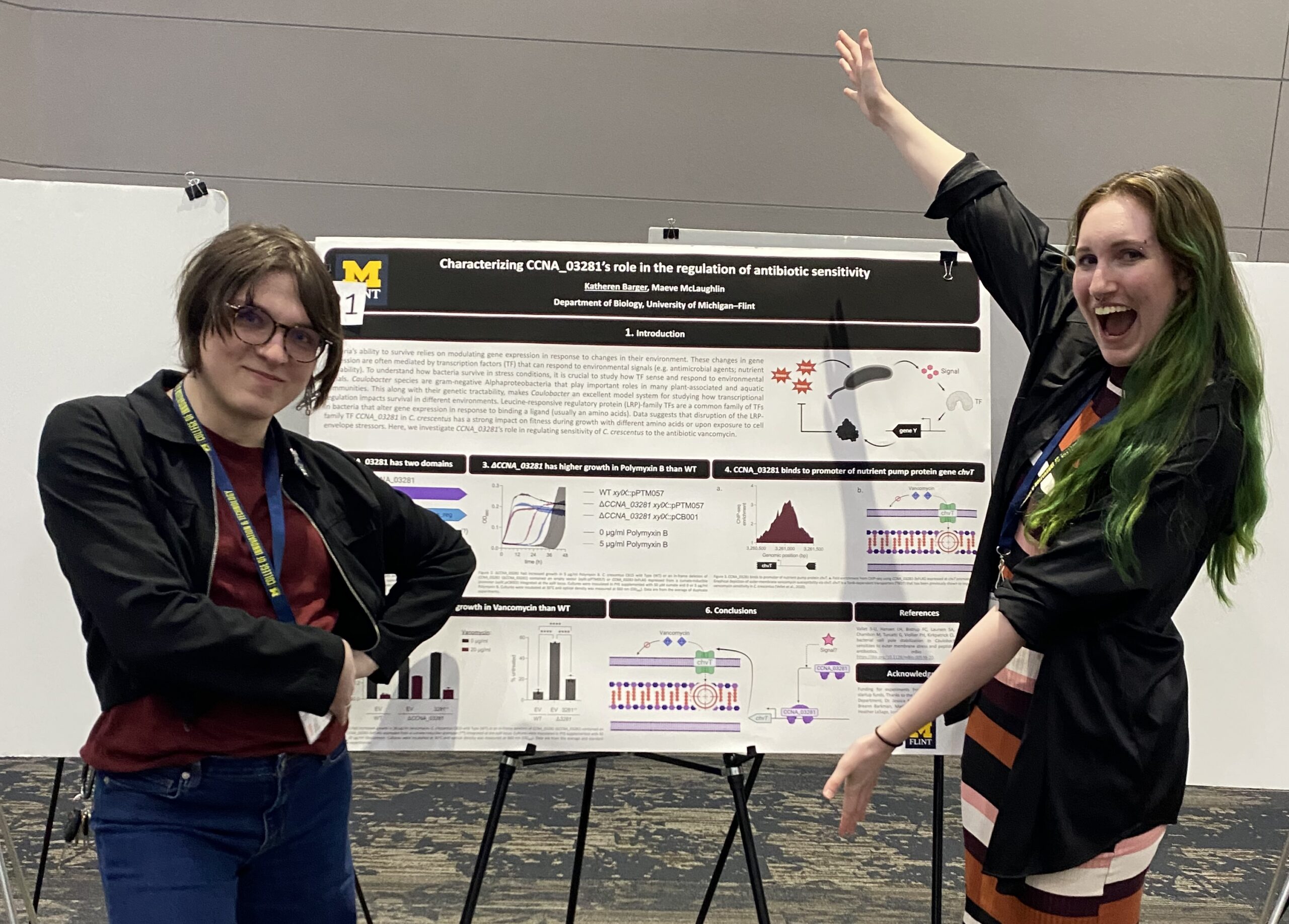 A professor and a student stand on either side of a research poster displayed on an easel at a conference. The poster displays charts, diagrams, and molecular illustrations related to bacterial antibiotic sensitivity research. The student on the right raises one arm and smiles broadly, while the professor on the left stands with arms crossed, smiling.