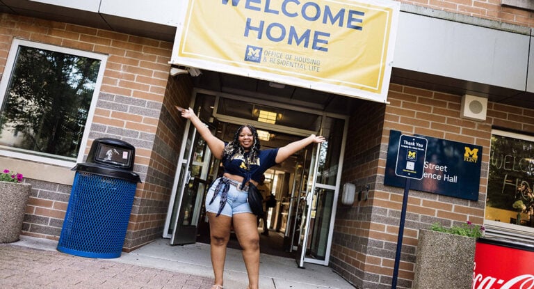 A person stands in front of a building entrance with arms raised, smiling. A large banner above reads "WELCOME HOME."