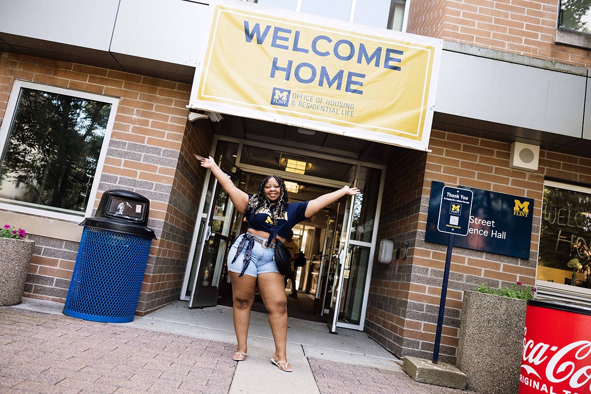 A person stands in front of a building entrance with arms raised, smiling. A large banner above reads "WELCOME HOME."