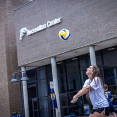 A young woman in a gray t-shirt and black shorts is preparing to hit a volleyball outside a building labeled "Recreation Center."