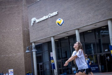 A young woman in a gray t-shirt and black shorts is preparing to hit a volleyball outside a building labeled "Recreation Center."