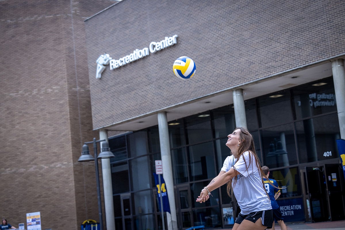 A young woman in a gray t-shirt and black shorts is preparing to hit a volleyball outside a building labeled "Recreation Center."