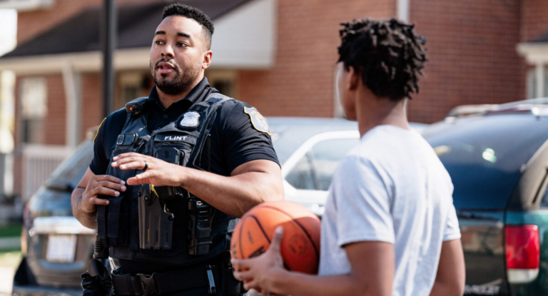 A police officer in a black uniform talks to a young person holding a basketball in a residential area.