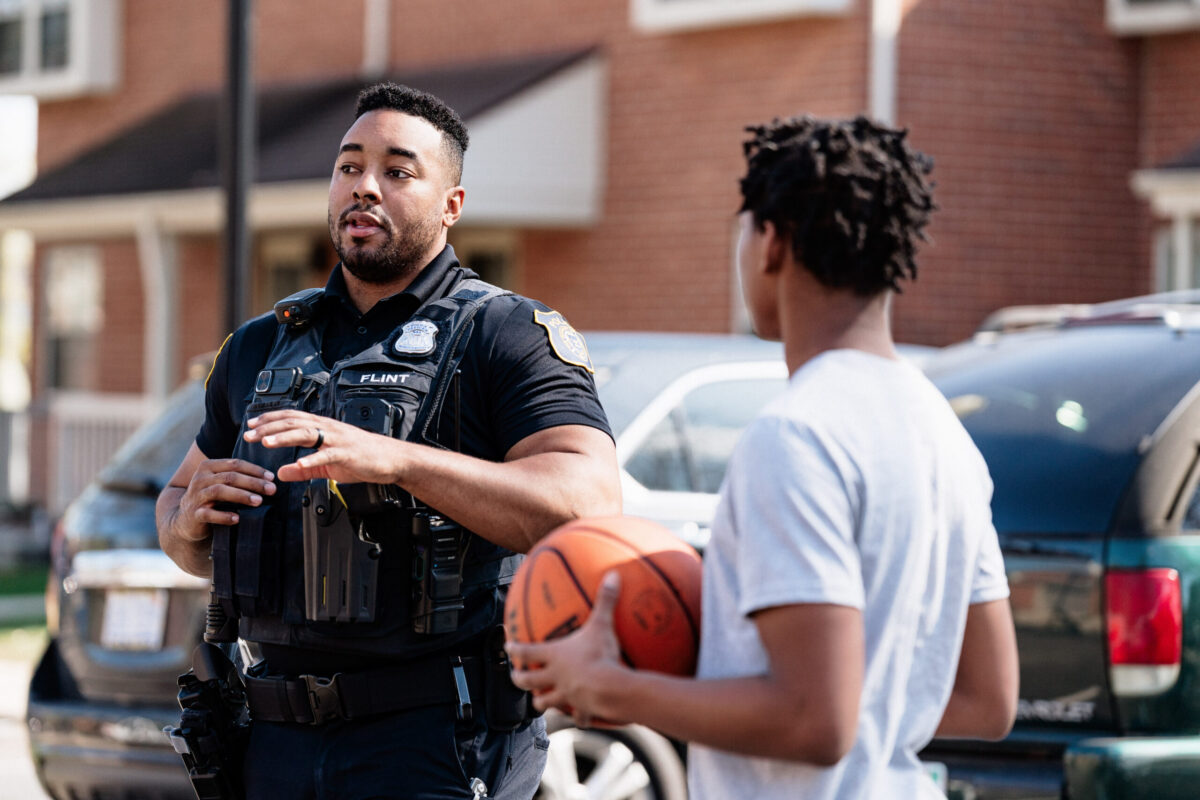 A police officer in a black uniform talks to a young person holding a basketball in a residential area.