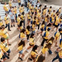 A large group of people wearing yellow shirts and blue backpacks walking together on a tiled outdoor area.