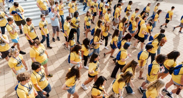 A large group of people wearing yellow shirts and blue backpacks walking together on a tiled outdoor area.