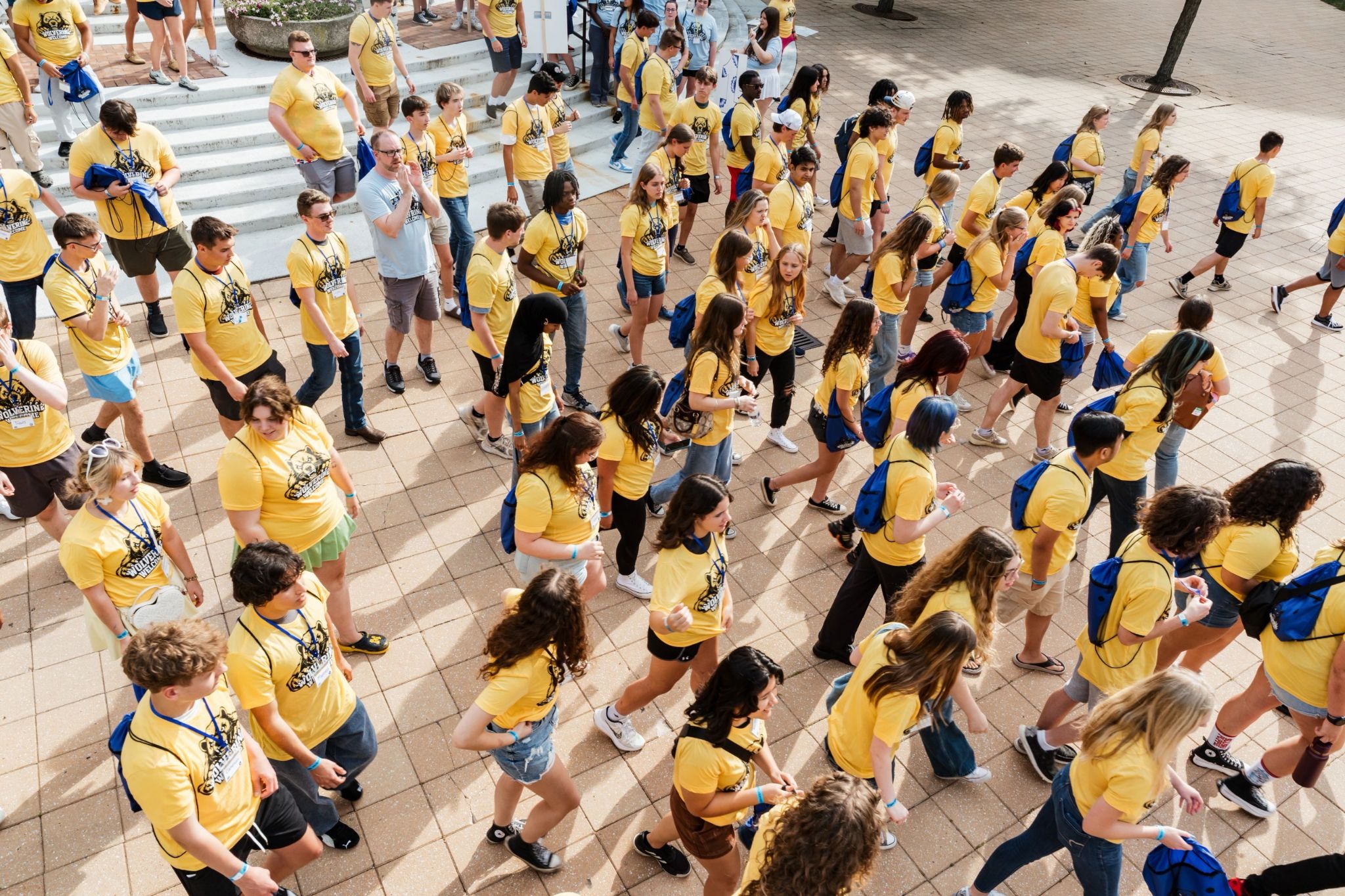 A large group of people wearing yellow shirts and blue backpacks walking together on a tiled outdoor area.