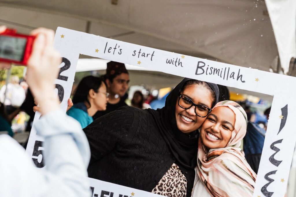 Two women smiling and posing together inside a decorative frame that says "let's start with Bismillah."