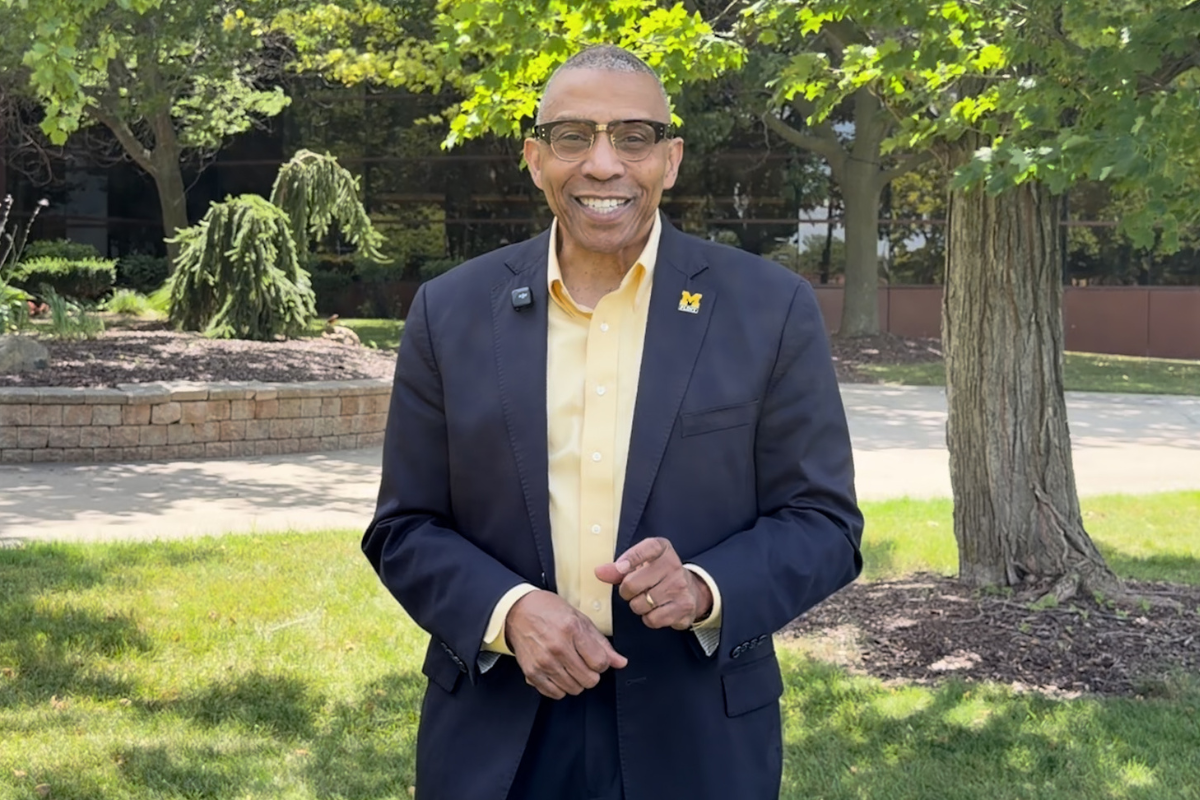 A smiling man wearing glasses and a dark suit stands outdoors in front of greenery, with a yellow shirt and a UM-Flint pin on his lapel.