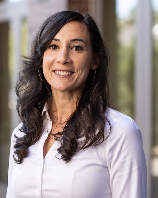 A woman with long, dark hair and hoop earrings smiles at the camera, wearing a white shirt, with a blurred background.