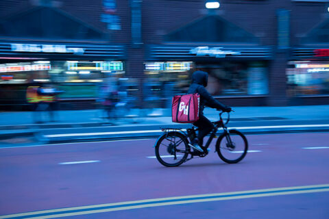 A person in a black hoodie rides a bicycle on a city street, carrying a large red delivery bag on their back. The background is blurred, indicating motion.