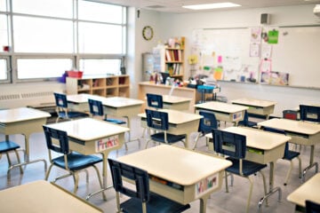 A classroom with several empty desks and chairs arranged in rows, a whiteboard and shelves in the background.