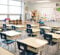 A classroom with several empty desks and chairs arranged in rows, a whiteboard and shelves in the background.