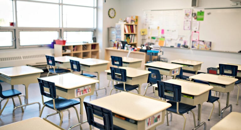 A classroom with several empty desks and chairs arranged in rows, a whiteboard and shelves in the background.
