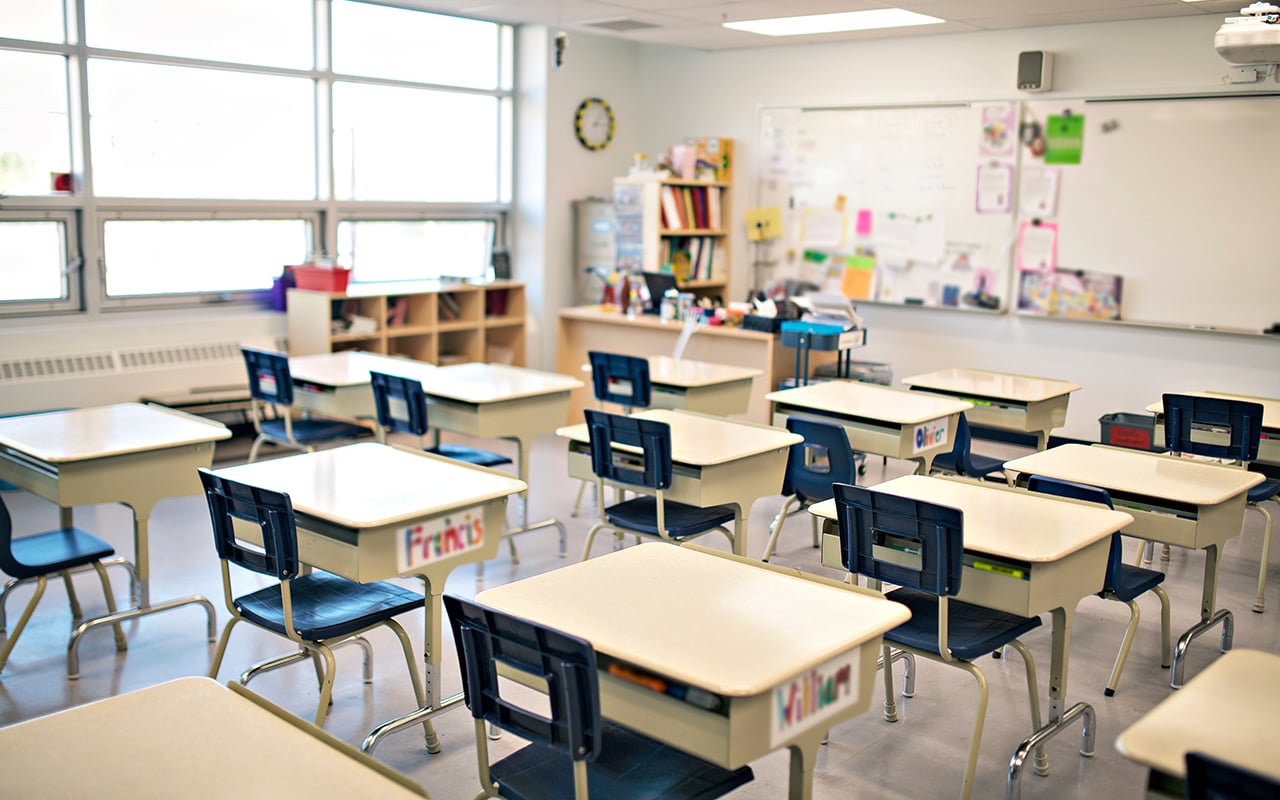 A classroom with several empty desks and chairs arranged in rows, a whiteboard and shelves in the background.