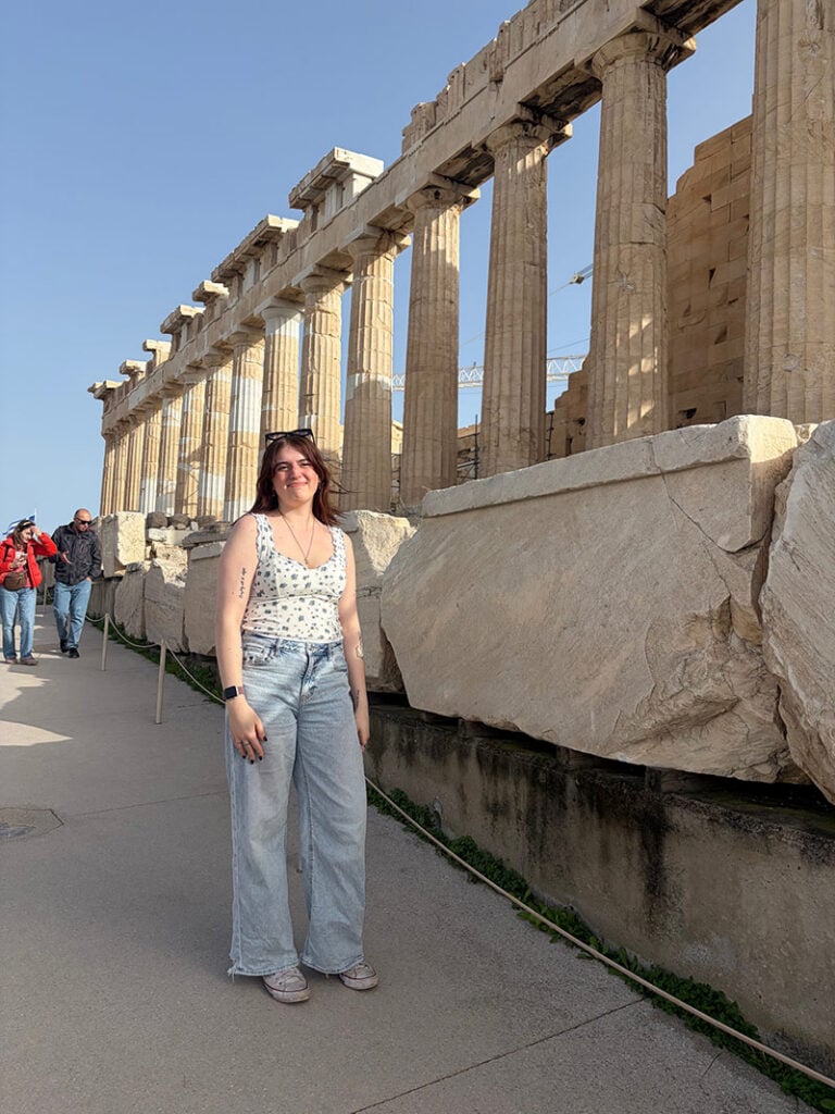A person stands in front of the Parthenon, a historic temple with tall columns, on a sunny day.
