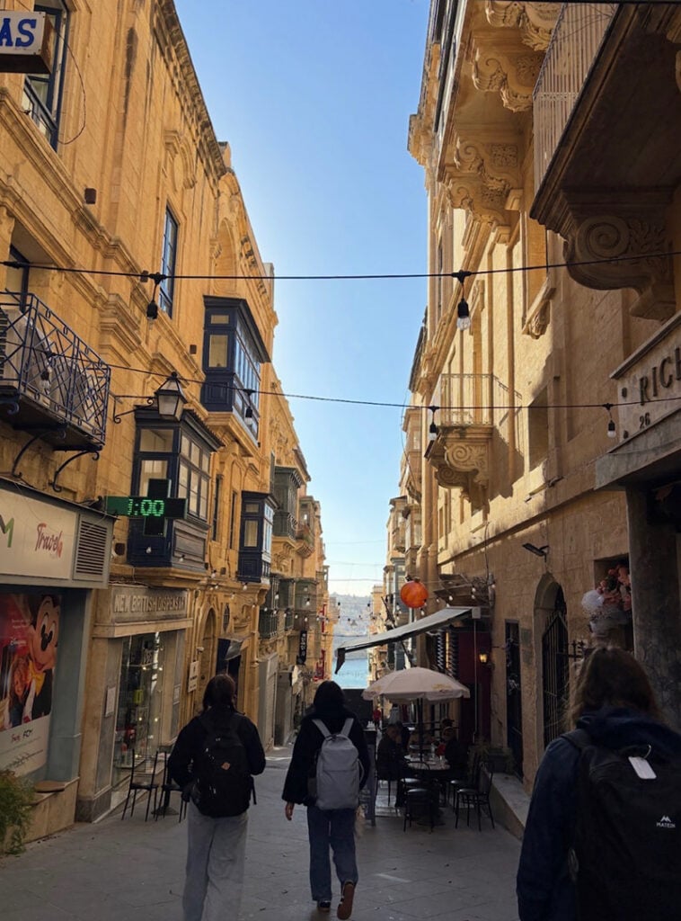A narrow street lined with historic buildings, featuring people walking towards a view of the sea in the distance. A digital clock displays "1:00" on a nearby wall.