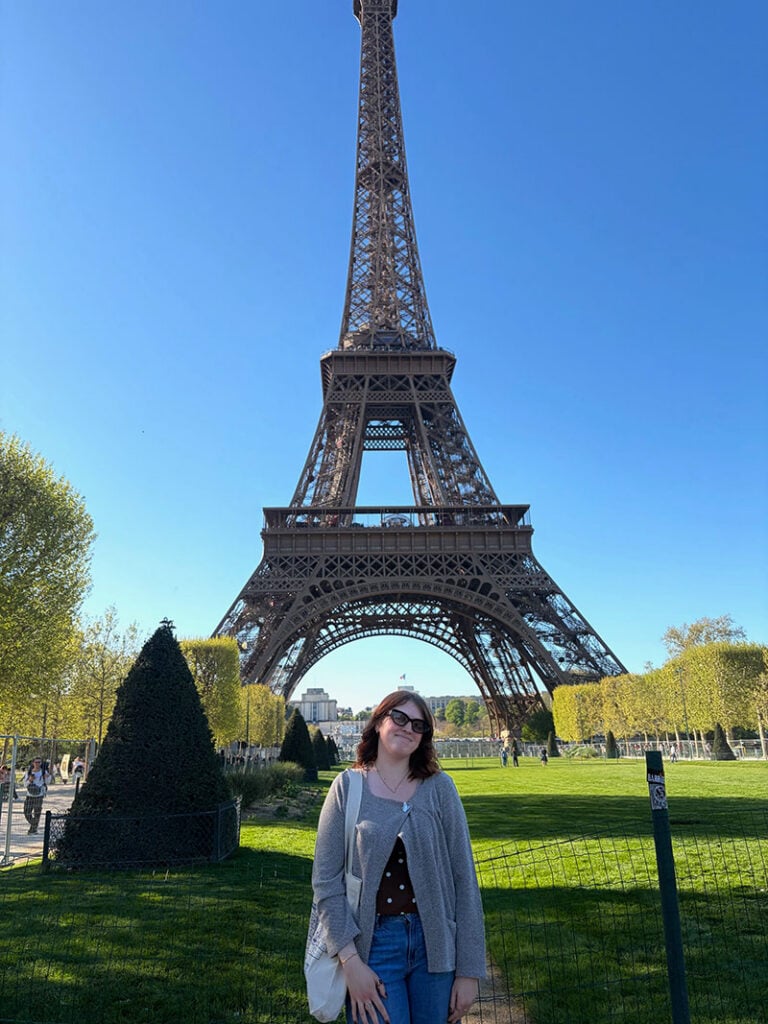 A person stands in front of the Eiffel Tower on a sunny day, surrounded by green grass and trees.