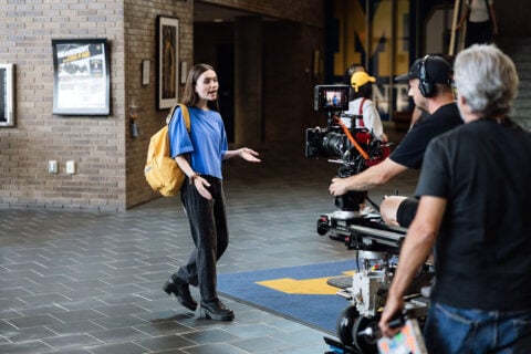 A young woman in a blue shirt and black pants is speaking animatedly in a hallway, while a film crew records her with a camera on a dolly. She has a yellow backpack and stands on a patterned floor.