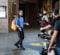 A young woman in a blue shirt and black pants is speaking animatedly in a hallway, while a film crew records her with a camera on a dolly. She has a yellow backpack and stands on a patterned floor.