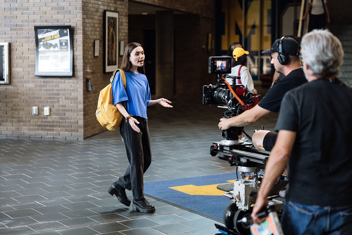 A young woman in a blue shirt and black pants is speaking animatedly in a hallway, while a film crew records her with a camera on a dolly. She has a yellow backpack and stands on a patterned floor.