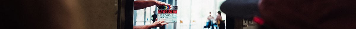 FLINT_BTS Commercial 2026_0009 A person is holding a clapperboard in front of a monitor displaying a scene being filmed, with a blurred background of people.