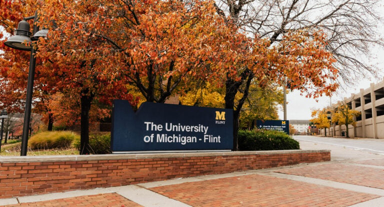 Sign for the University of Michigan-Flint, surrounded by autumn foliage, with a brick pathway in the foreground.