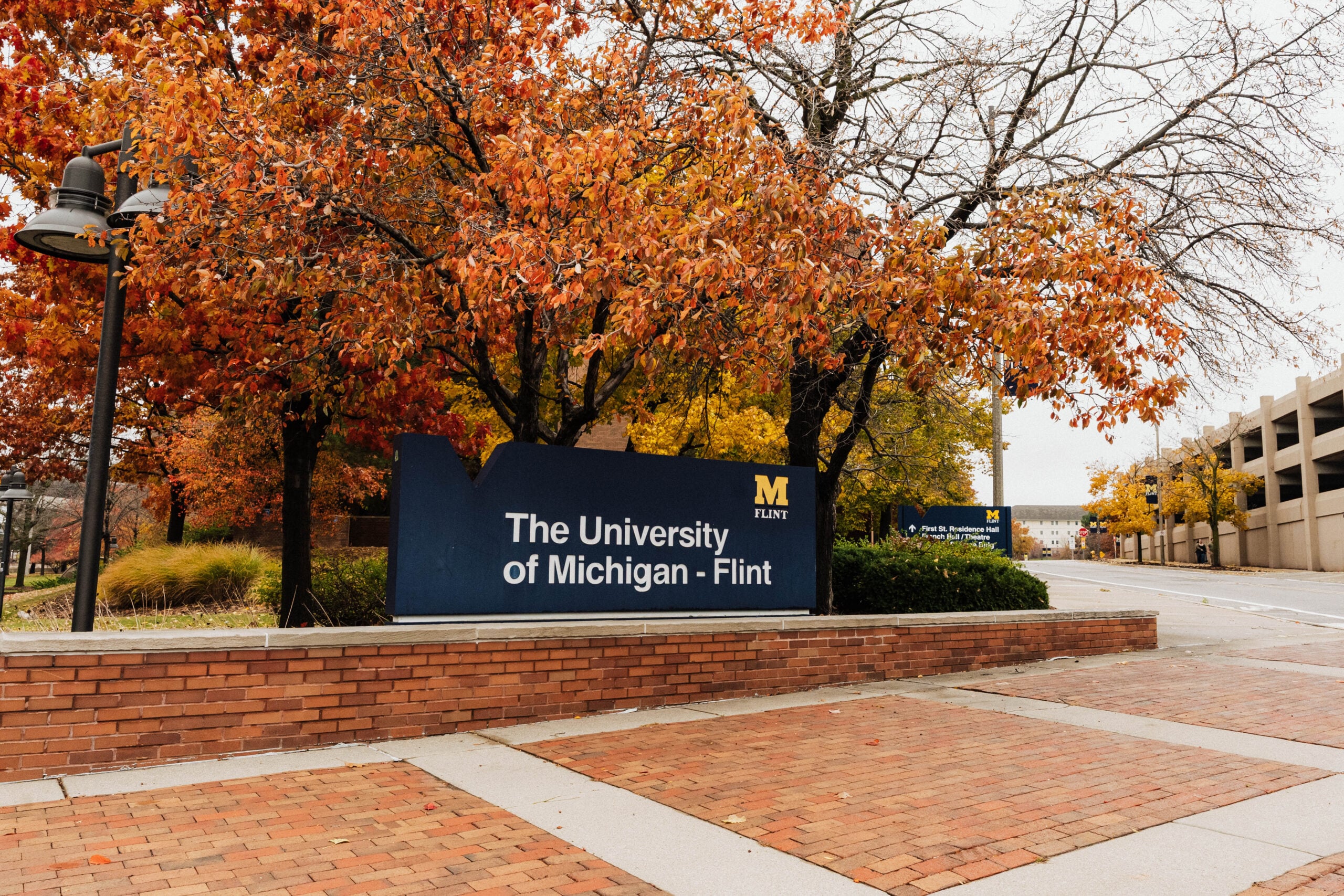 Sign for the University of Michigan-Flint, surrounded by autumn foliage, with a brick pathway in the foreground.