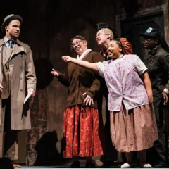 A scene from a theatrical performance featuring five actors. A man in a trench coat stands to the left, while four women in vintage clothing interact with him, expressing excitement. A vintage table with a green object is visible in the background.