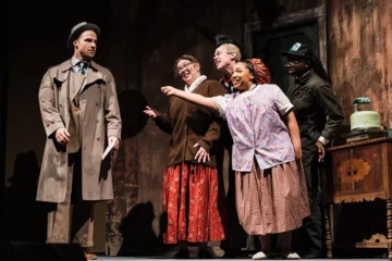 A scene from a theatrical performance featuring five actors. A man in a trench coat stands to the left, while four women in vintage clothing interact with him, expressing excitement. A vintage table with a green object is visible in the background.