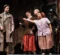 A scene from a theatrical performance featuring five actors. A man in a trench coat stands to the left, while four women in vintage clothing interact with him, expressing excitement. A vintage table with a green object is visible in the background.