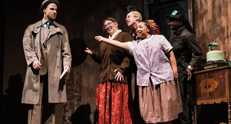 A scene from a theatrical performance featuring five actors. A man in a trench coat stands to the left, while four women in vintage clothing interact with him, expressing excitement. A vintage table with a green object is visible in the background.