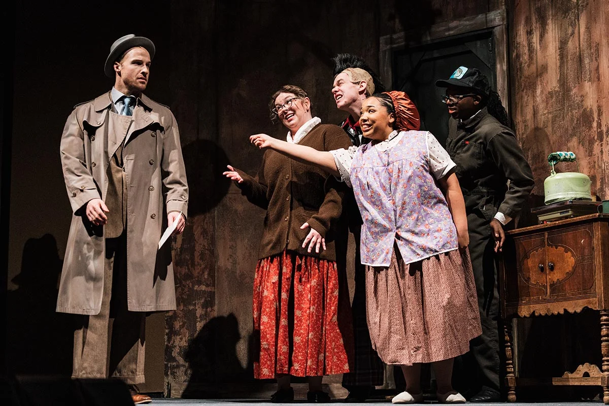 A scene from a theatrical performance featuring five actors. A man in a trench coat stands to the left, while four women in vintage clothing interact with him, expressing excitement. A vintage table with a green object is visible in the background.