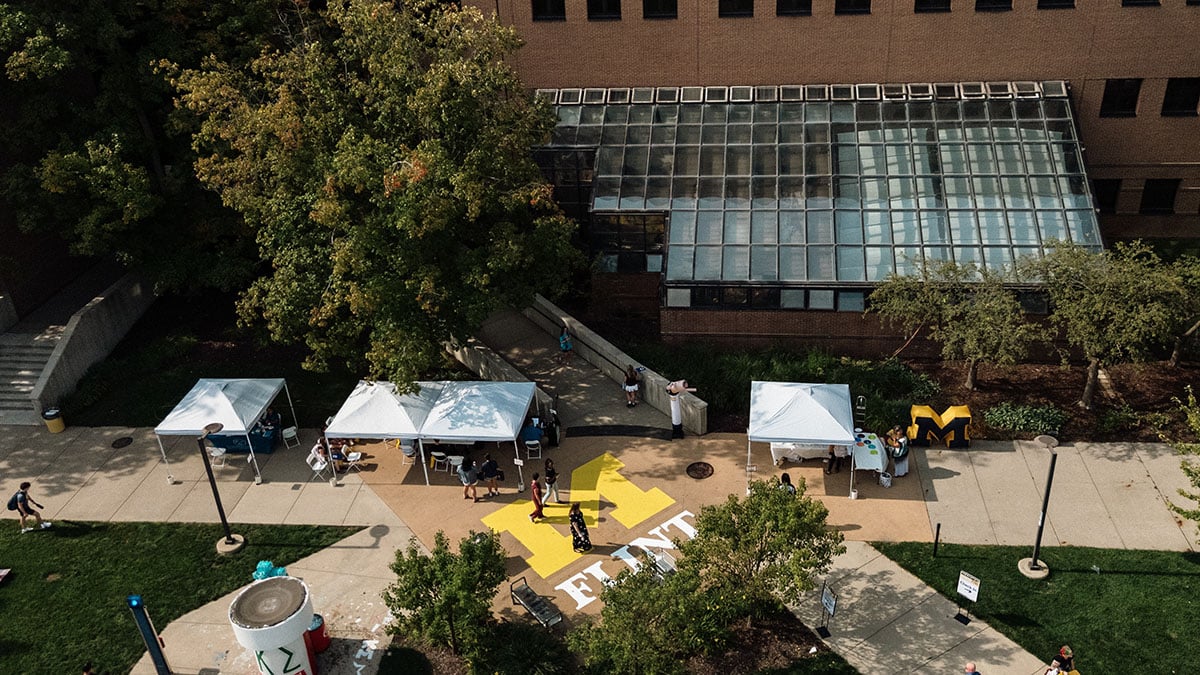 default An aerial view of a campus area featuring several white tents, people gathered around, and a large yellow "FLINT" painted on the ground. In the background, there is a glass building and trees.