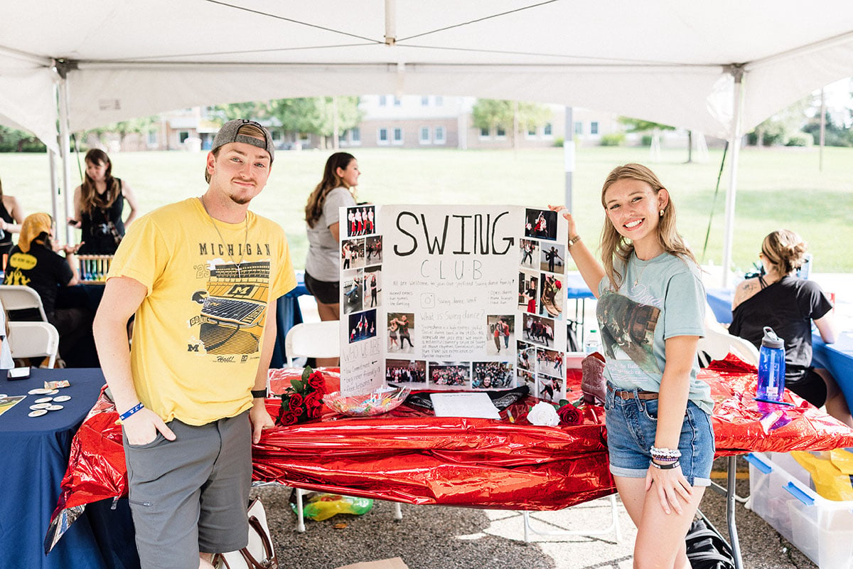 FLINT_M'Gagement Fair 2026_0007 A young man in a yellow shirt and a young woman in a blue shirt stand beside a display board for the "SWING CLUB" at an outdoor event. The board features photos and information about the club.