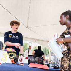 A young man in a Baja Racing shirt is speaking to a woman holding a water bottle and papers at a booth under a tent. A helmet and number "163" are on the table.