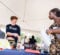A young man in a Baja Racing shirt is speaking to a woman holding a water bottle and papers at a booth under a tent. A helmet and number "163" are on the table.