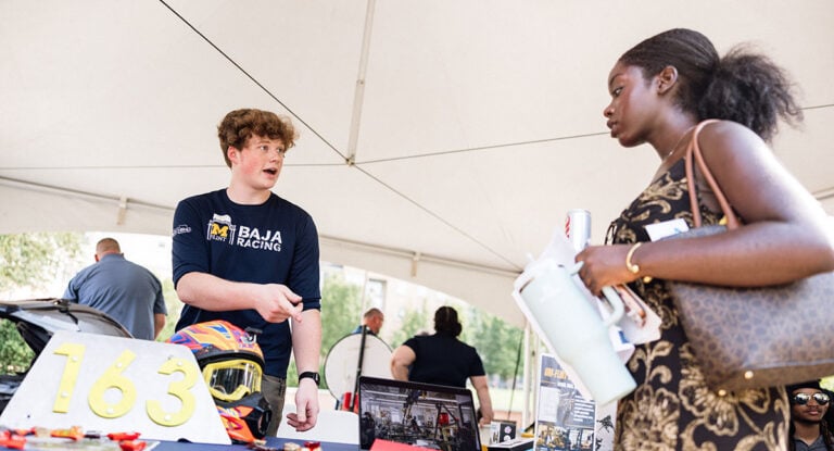 A young man in a Baja Racing shirt is speaking to a woman holding a water bottle and papers at a booth under a tent. A helmet and number "163" are on the table.