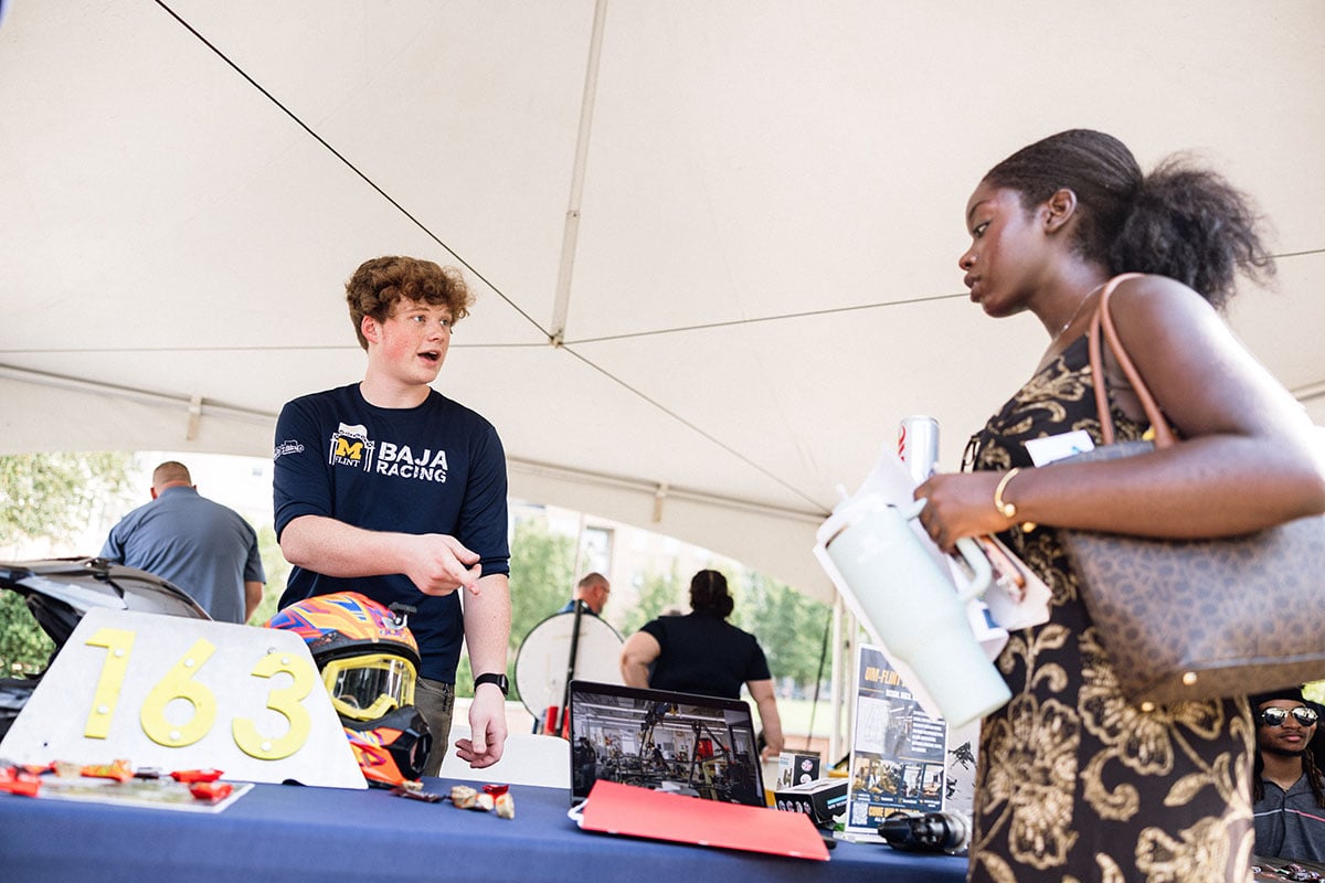 A young man in a Baja Racing shirt is speaking to a woman holding a water bottle and papers at a booth under a tent. A helmet and number "163" are on the table.