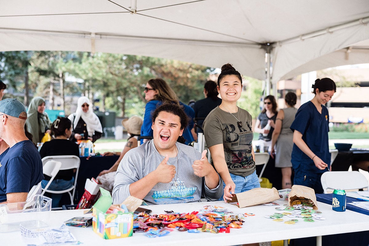 FLINT_M'Gagement Fair 2026_0014 Two young adults are smiling and giving thumbs up at a table filled with snacks and drinks, under a tent at an outdoor event.