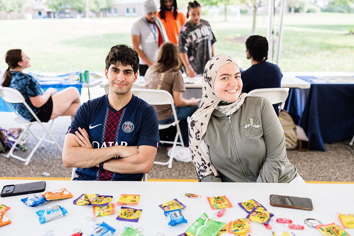 FLINT_M'Gagement Fair 2026_0017 A young man in a Paris Saint-Germain jersey and a young woman in a green jacket and hijab smile at the camera, sitting at a table with colorful snacks and keychains. In the background, other people are engaged in activities under a tent.