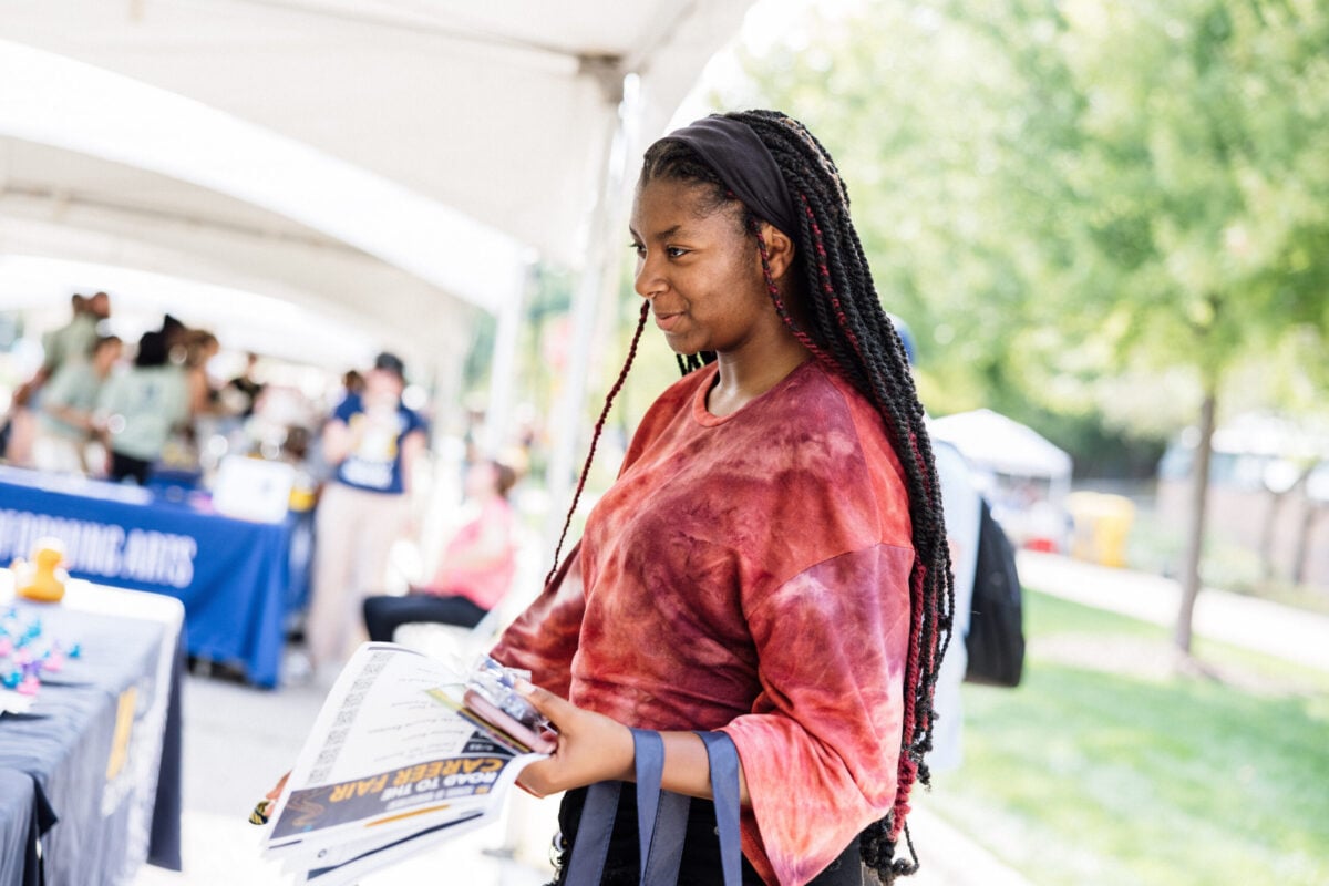 FLINT_M'Gagement Fair 2026_0019 A young woman with long braided hair is holding a stack of papers while walking through an outdoor event with tents.