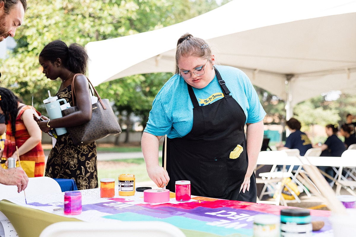 FLINT_M'Gagement Fair 2026_0021 A person with glasses and a blue shirt is painting on a colorful tablecloth at an outdoor event, with others in the background.